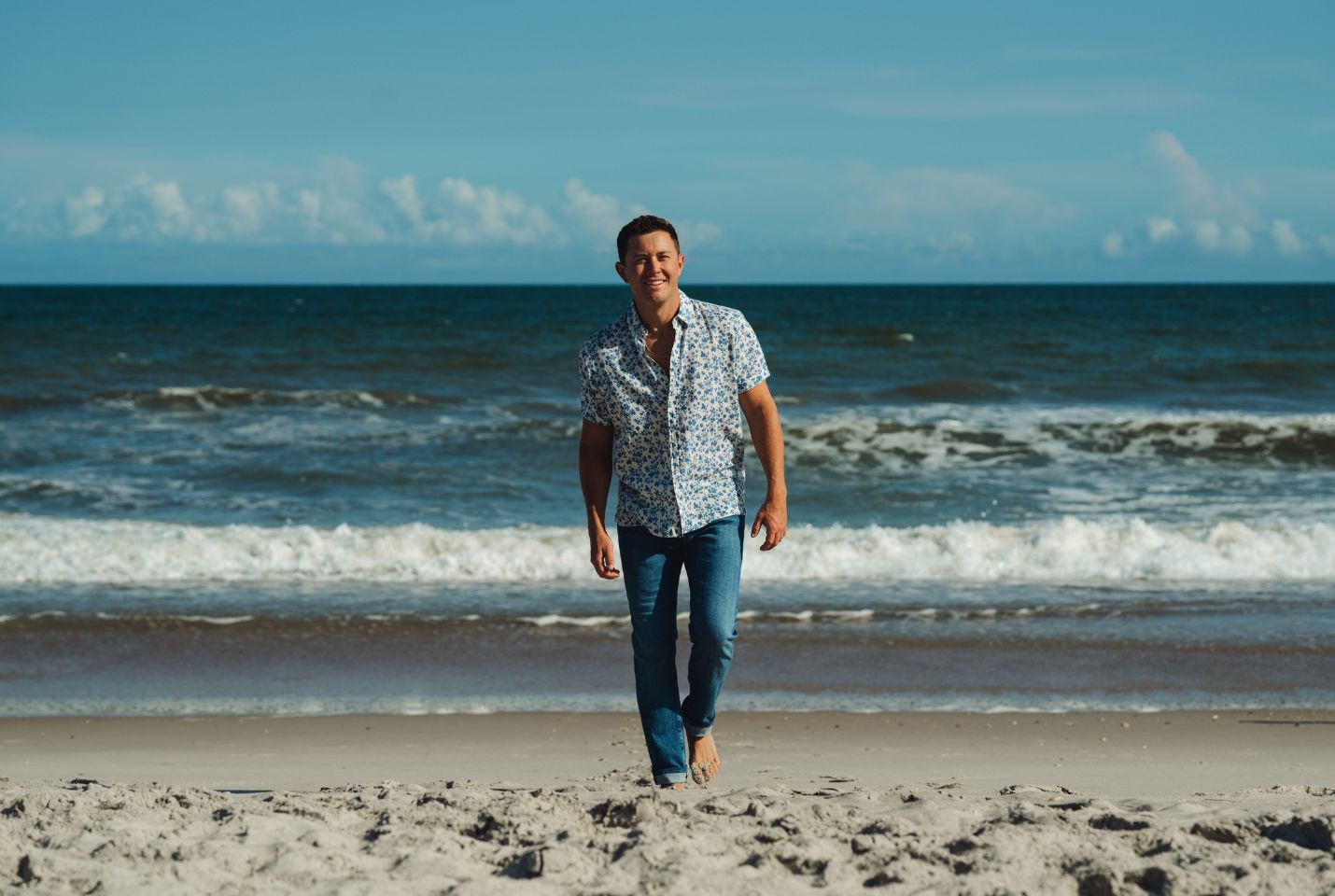 A man walking away from the sea on a beach wearing a blue printed shirt and blue jeans
